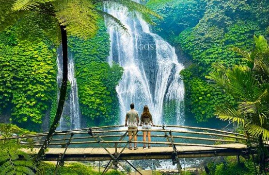 A couple of travelers standing hand-in-hand on a rustic bamboo footbridge, looking out at the spectacular streams of Banyumala Waterfall.