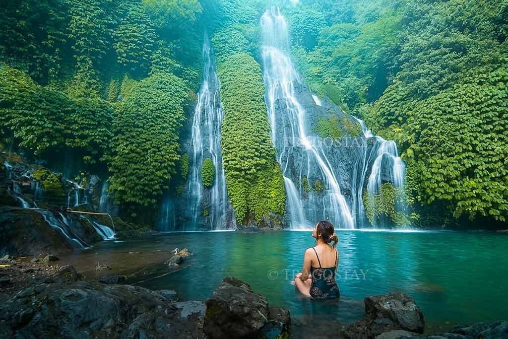 A female traveler relaxing and soaking in the cool, clear water of the plunge pool at Banyumala Waterfall while enjoying the peaceful nature.