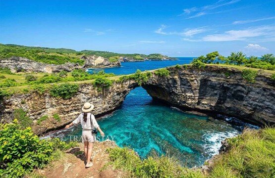 A female tourist standing on the cliff edge facing the iconic natural archway of Broken Beach on a sunny day.