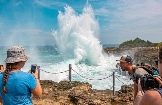 Tourists using cameras and phones to capture the dramatic wave splashes at Devil’s Tears under a clear blue sky.