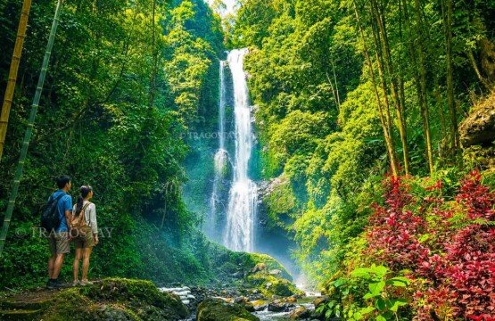A male and female tourist standing side-by-side on a rocky path, looking up in awe at the tall rushing waters of Melanting Waterfall.