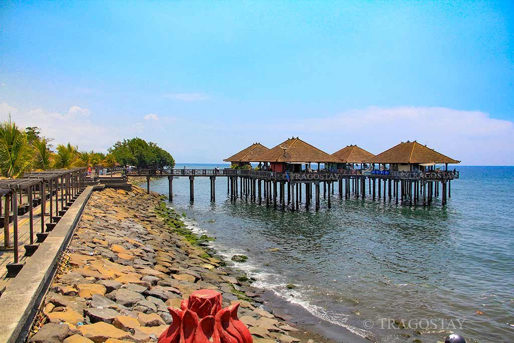A sunny daytime view of Ex-Buleleng Harbour showing several seafood restaurants built on stilts directly over the calm blue sea.