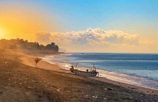 A peaceful sunrise over Goa Lawah Beach featuring a traditional outrigger boat (jukung) resting on the black sand.
