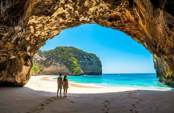 A tourist couple standing inside a natural cliff cave looking out at the sunny ocean at Kelingking Beach.