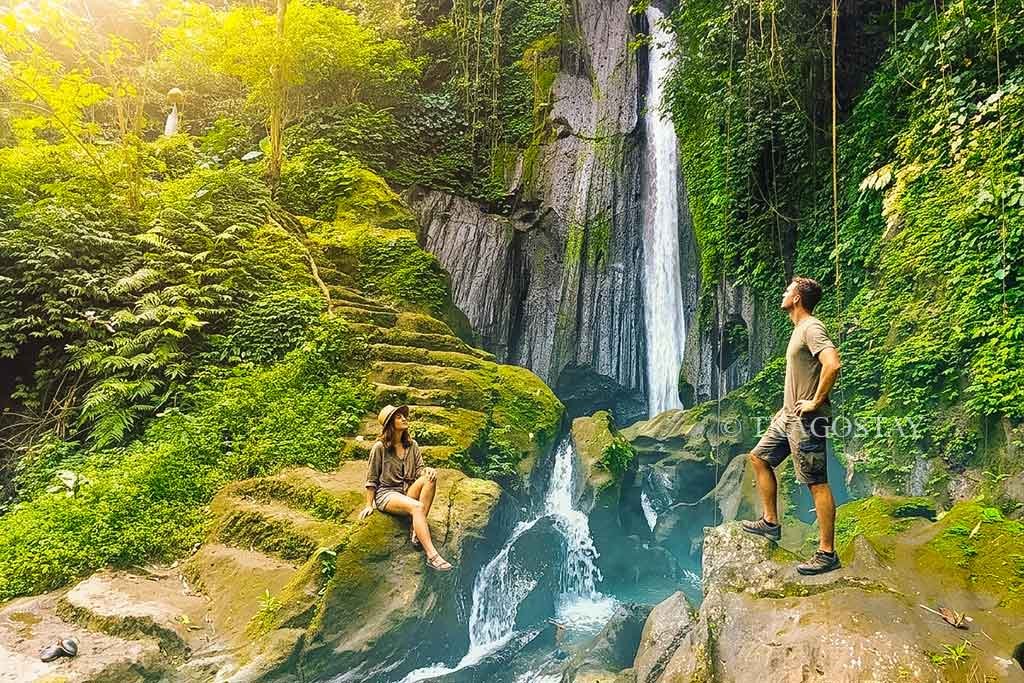 A traveler couple standing on the riverbank posing together with the tall Kuning Waterfall in the background.