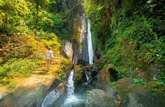 A woman looking up in awe at the massive height and sheer drop of Kuning Waterfall from the rocky base.