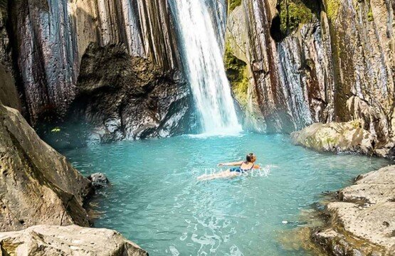 A woman swimming peacefully in the clear, natural rock plunge pool at the base of Kuning Waterfall.