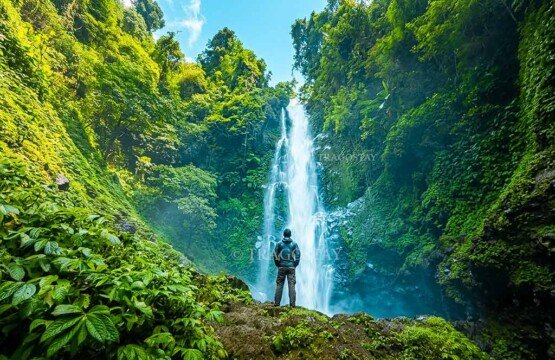 A lone traveler standing on a small grassy earth mound, looking out toward the magnificent tall white cascades of Melanting Waterfall.