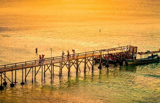 A scenic evening view of a rustic wooden jetty stretching into the calm sea at Menjangan Island during a spectacular golden sunset.
