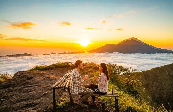 A couple of travelers drinking hot coffee at the top of Mount Batur while basking in the warm glow of a clear sunrise.