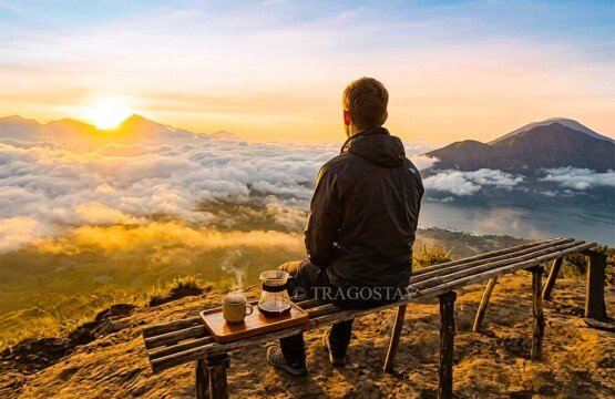 A man sitting calmly on a wooden bench looking out at the sunrise with a hot cup of coffee by his side on Mount Batur.