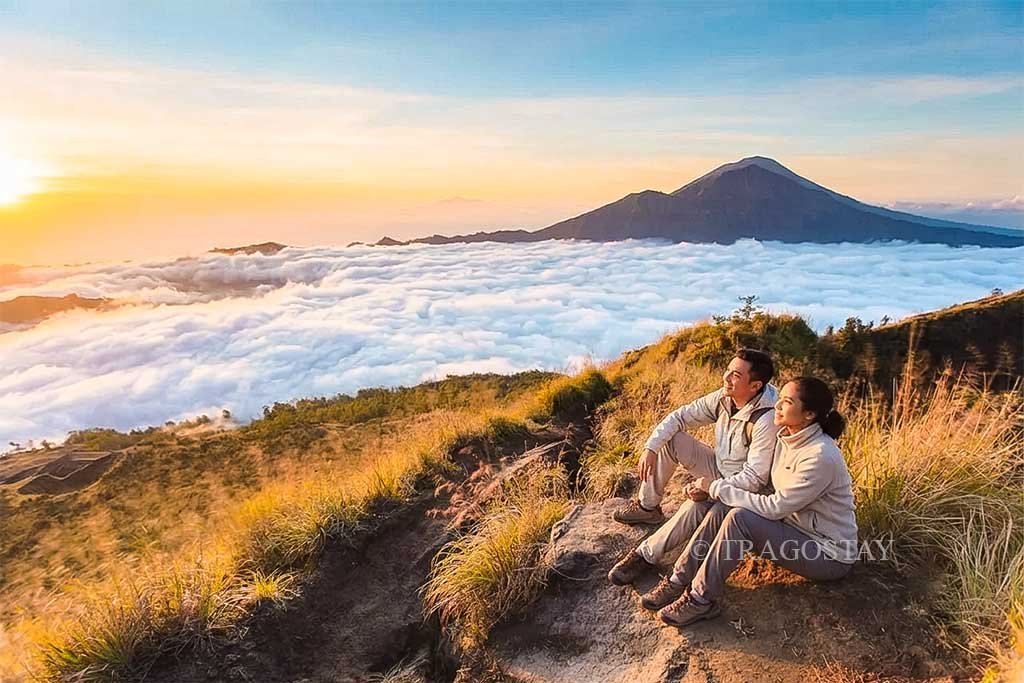 A tourist couple sitting together on the rocky peak of Mount Batur, enjoying a bright sunrise above the low clouds.