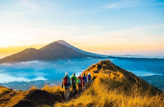A group of international tourists hiking along the narrow dirt pathway on the ridge of Mount Batur during a golden sunrise.