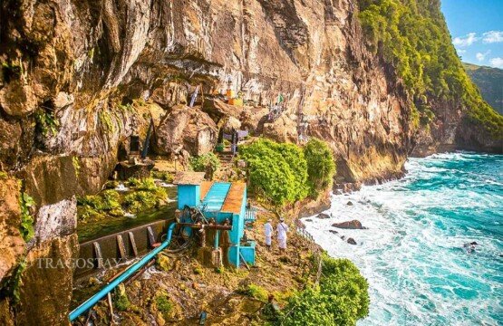 Two Balinese people in traditional attire walking on coastal rocks toward the sacred temple at Peguyangan Waterfall.