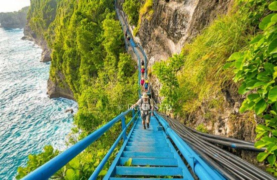 International tourists carefully climbing the steep blue iron stairs bolted to the cliff at Peguyangan Waterfall.