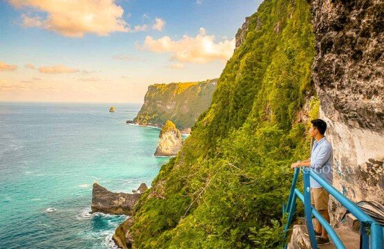 A man standing on a narrow cliffside path at Peguyangan Waterfall enjoying the panoramic view of the blue ocean.
