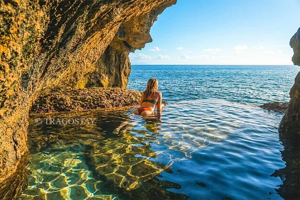 A female tourist soaking in a natural rock pool overlooking the ocean at Peguyangan Waterfall during the day.
