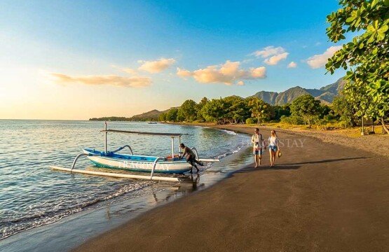 A tourist couple walking hand-in-hand along the quiet shore of Pemuteran Beach during a bright, silent morning.