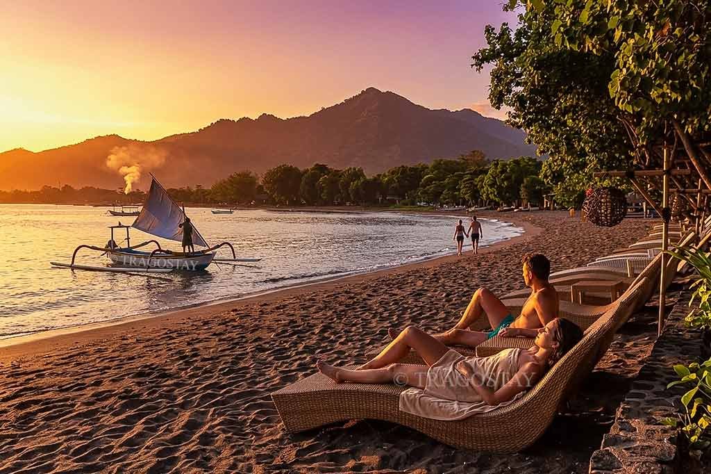 A tourist couple relaxing side-by-side on comfortable wooden sun beds while enjoying the morning scenery at Pemuteran Beach.