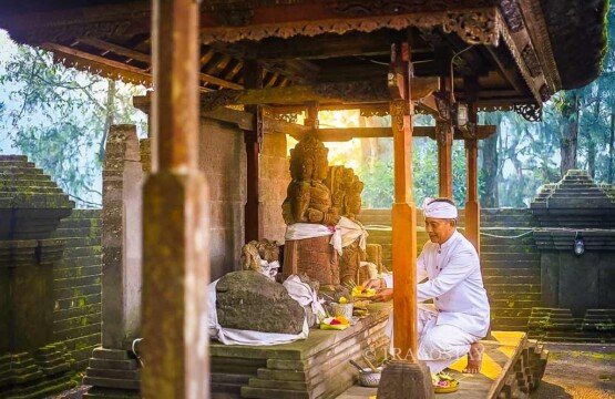 A Balinese man in traditional sarong placing a Canang Sari offering in front of a weathered ancient stone statue at Puncak Penulisan Temple.