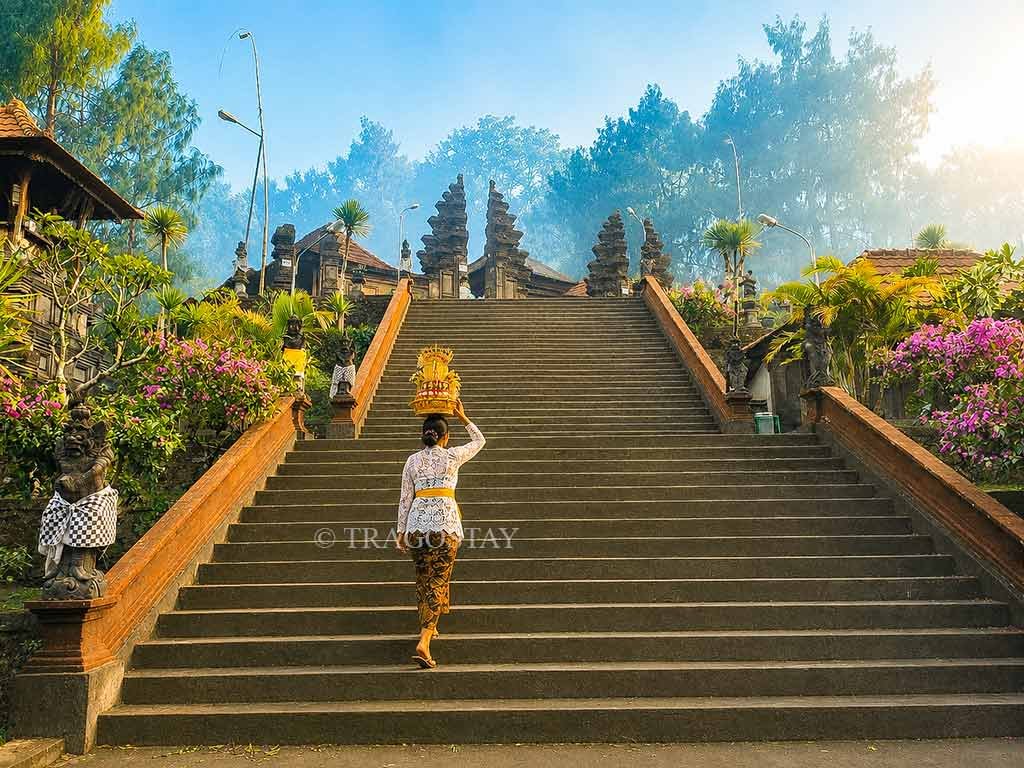 A Balinese woman in ceremonial attire balancing a tall Gebogan fruit tower on her head while climbing the steep stairs of Puncak Penulisan Temple.