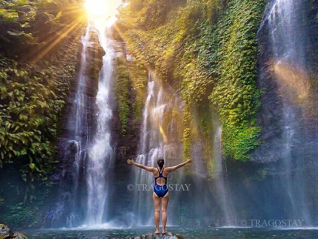 A female tourist standing with open arms, expressing joy and freedom on a river rock directly in front of the spraying Sekumpul Waterfall.