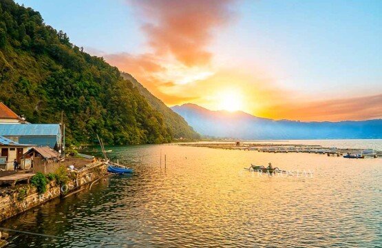 A vibrant sunset over the shore of Trunyan Village featuring a local fisherman rowing his canoe between floating fish farm cages.
