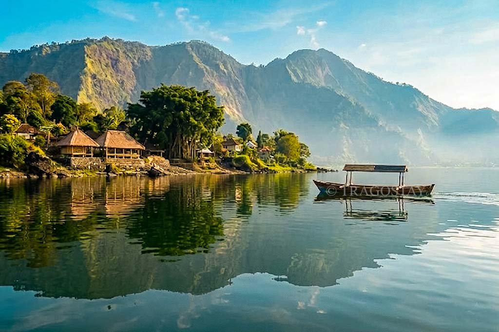 A peaceful morning landscape of smooth, glassy lake water with a traditional fishing boat and Trunyan Village visible along the sunlit coastline.