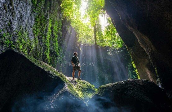 A man standing confidently on top of a large boulder inside the rocky cave chamber of Tukad Cepung Waterfall.