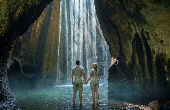 A tourist couple standing inside the cave canyon, admiring the powerful flow of Tukad Cepung Waterfall from a close distance.