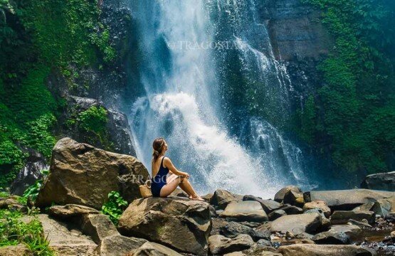 A female traveler sitting peacefully on a large river boulder, looking up in awe at the spectacular flow of Gitgit Waterfall.