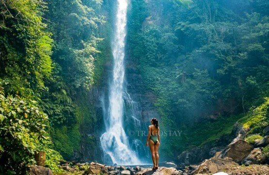 A female tourist standing on a large stone plate, completely captivated by the stunning beauty and cool mist of Gitgit Waterfall.