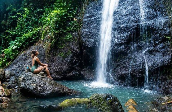 A female traveler sitting peacefully on a large rock while looking up at the top of Yeh Labuh Waterfall.