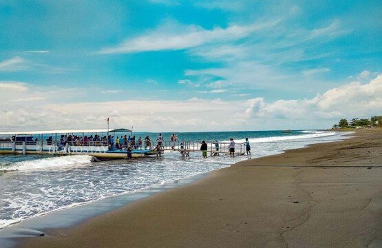 A busy scene at the Kusamba Beach pier showing passengers boarding and disembarking from a fast boat transport ferry.